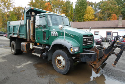 Truck front-left view of a green 2010 Mack GU Granite dump truck with a front-mounted plow/loader, showing Mack logo on grill and wheel arches, and rust around the wheel rims.
