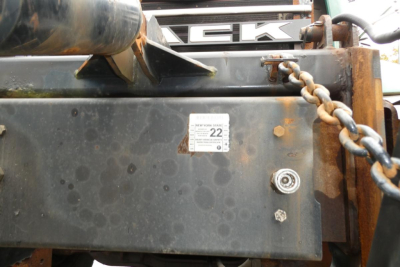 Truck front end showing Mack logo and attached chain with rust on metal panel; small white sticker with numbers; hydraulic hose visible.