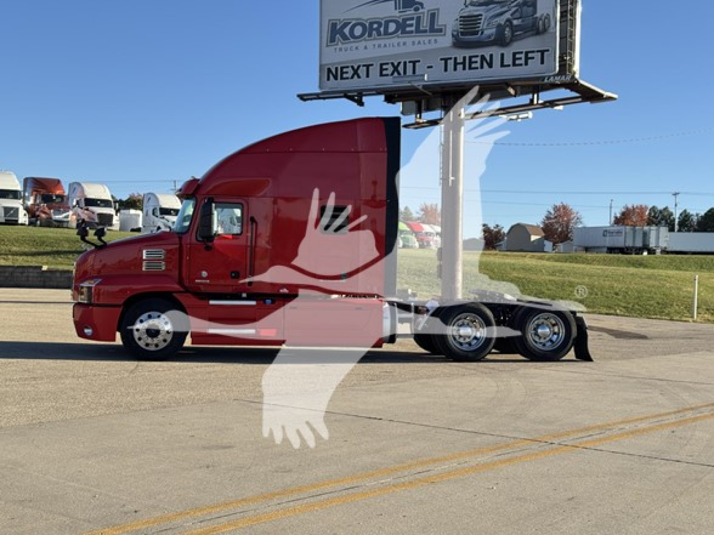 Truck cab and chassis showing Mack Anthem 64T with red cab and sleeper, side view parked above street with dealership sign in background.