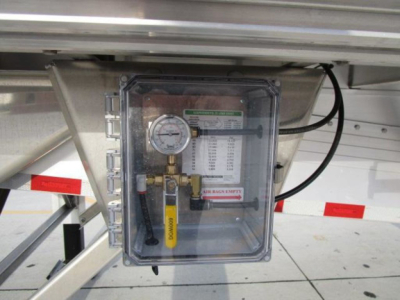 Control panel with pressure gauge and labeled warning on a metal box mounted under a flatbed trailer; valve with yellow handle, tubing, and wiring visible.