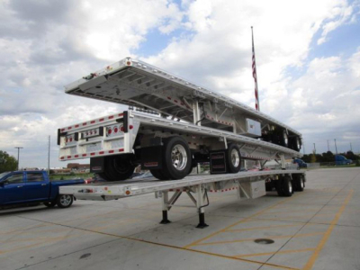 Flatbed trailer in raised stance showing dual drop-down sections and spare tire mounted on side with safety rails and reflective tape.