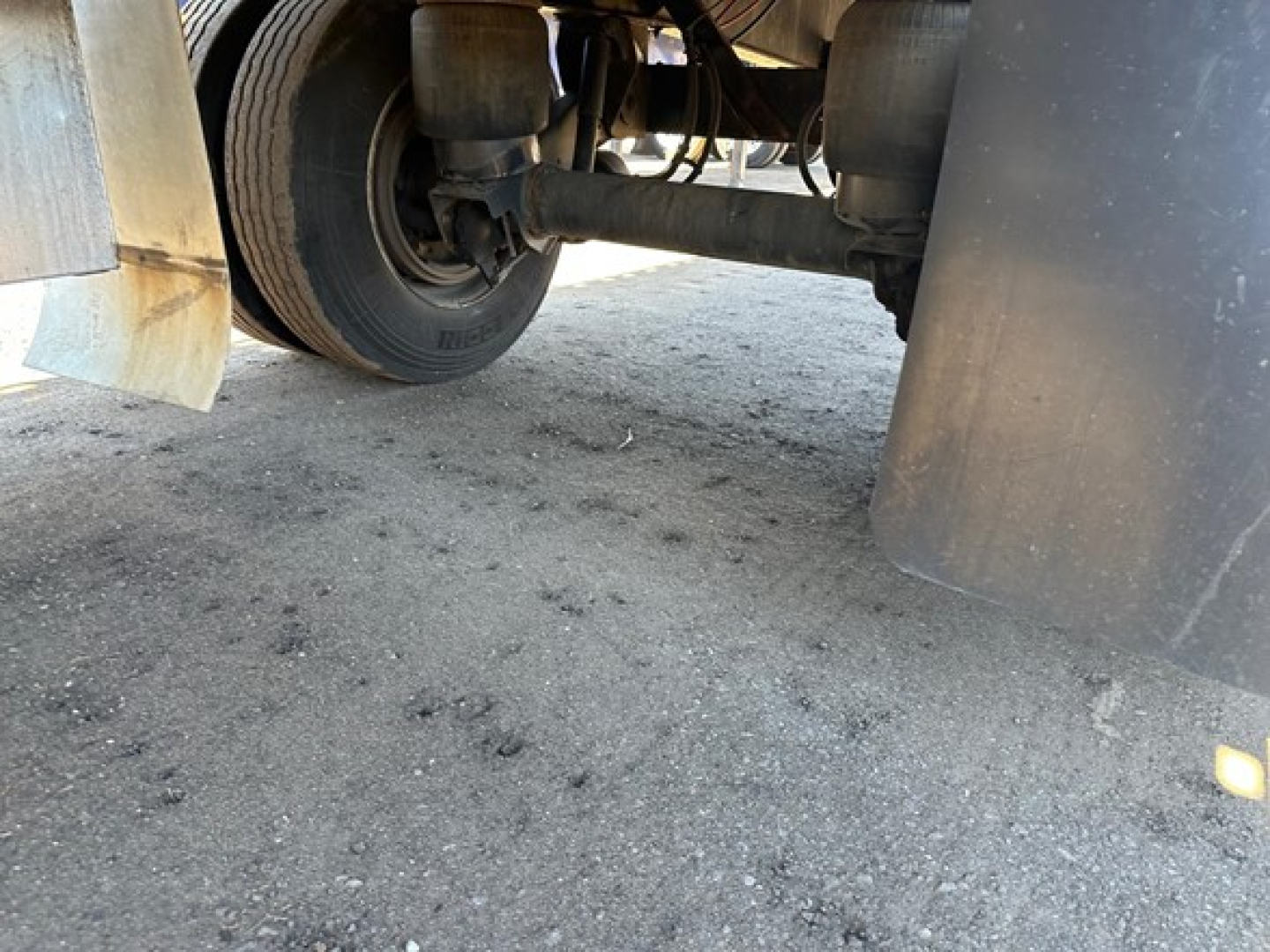 Trailer undercarriage showing wheel, axle, and mudflaps; dirt and gravel surface visible.
