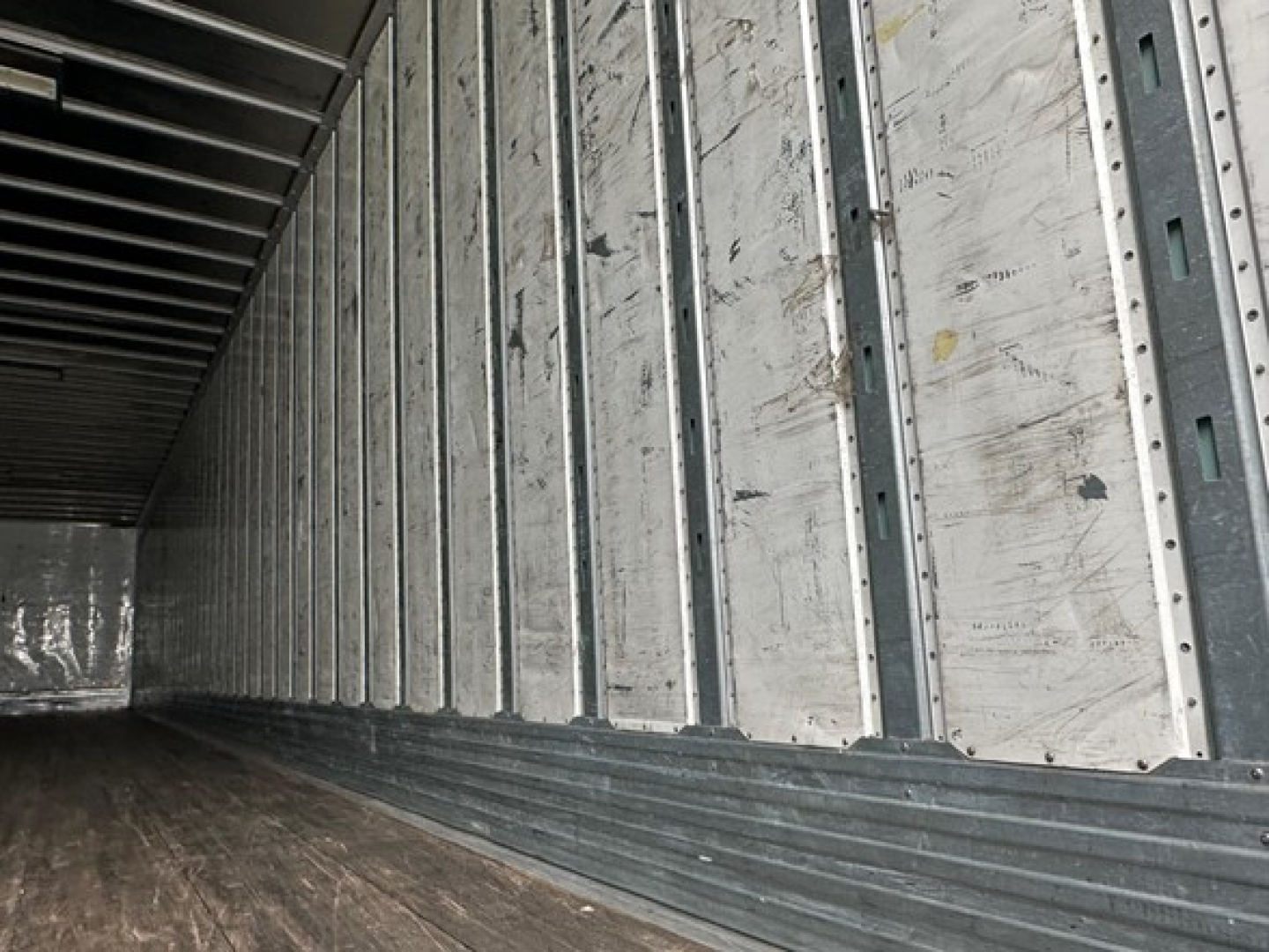 Trailer interior wall with vertical metal studs and ribbed floor showing worn aluminum panels and interior trailer space.