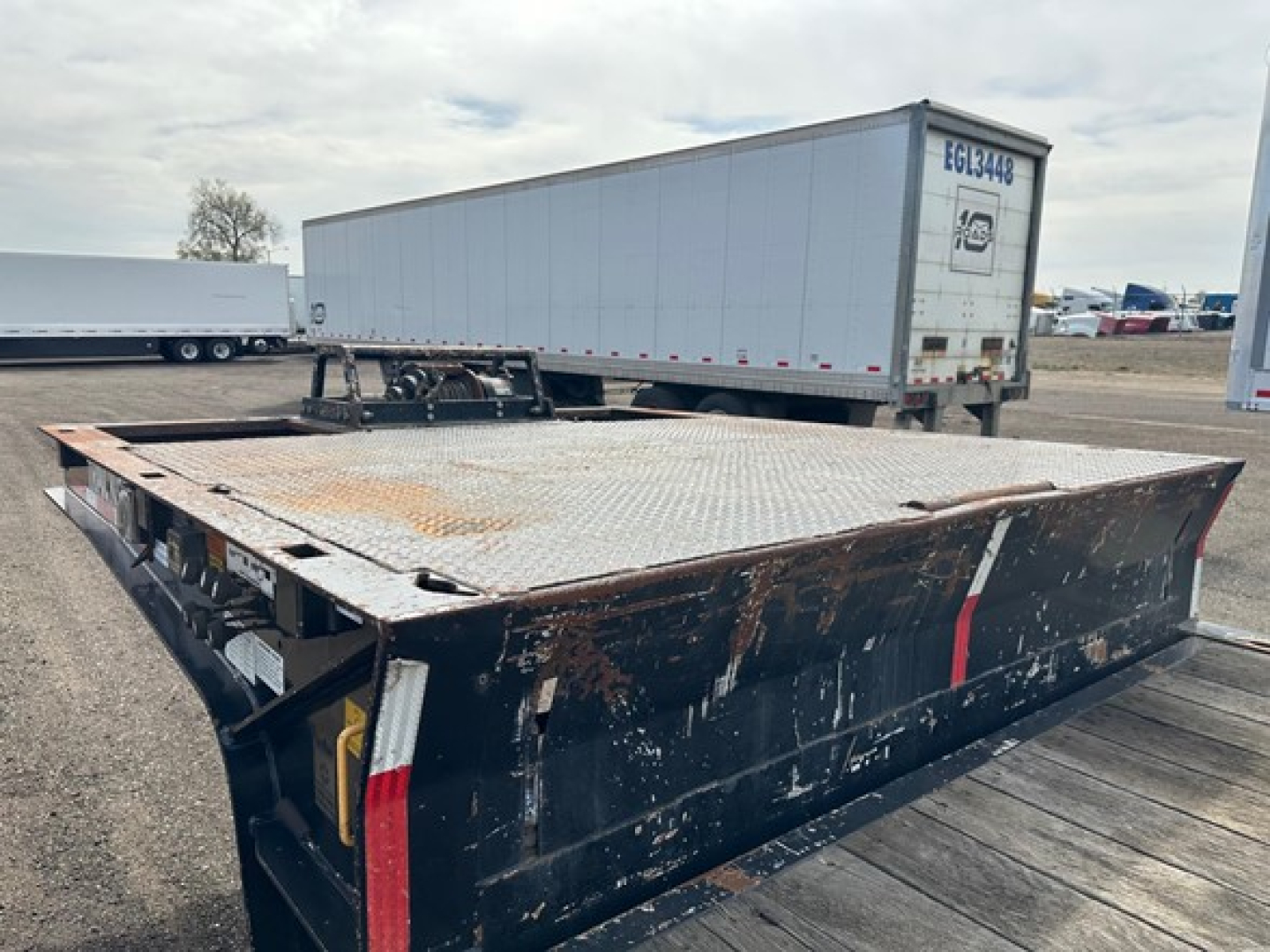 Trailer showing a large flatbed with a diamond plate deck and a black ramp; rust spots on deck edge; rear kingpin area visible; background includes semi trailers and a white dry van.