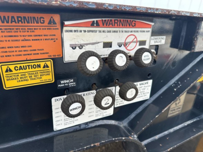 Trailer information panel showing warnings and control labels: three circular knobs labeled 1, 2, and 3, with adjacent warnings about WINCH, DO NOT OPERATE, and hydraulic ramp. Several warning decals in yellow and orange with text, and a di