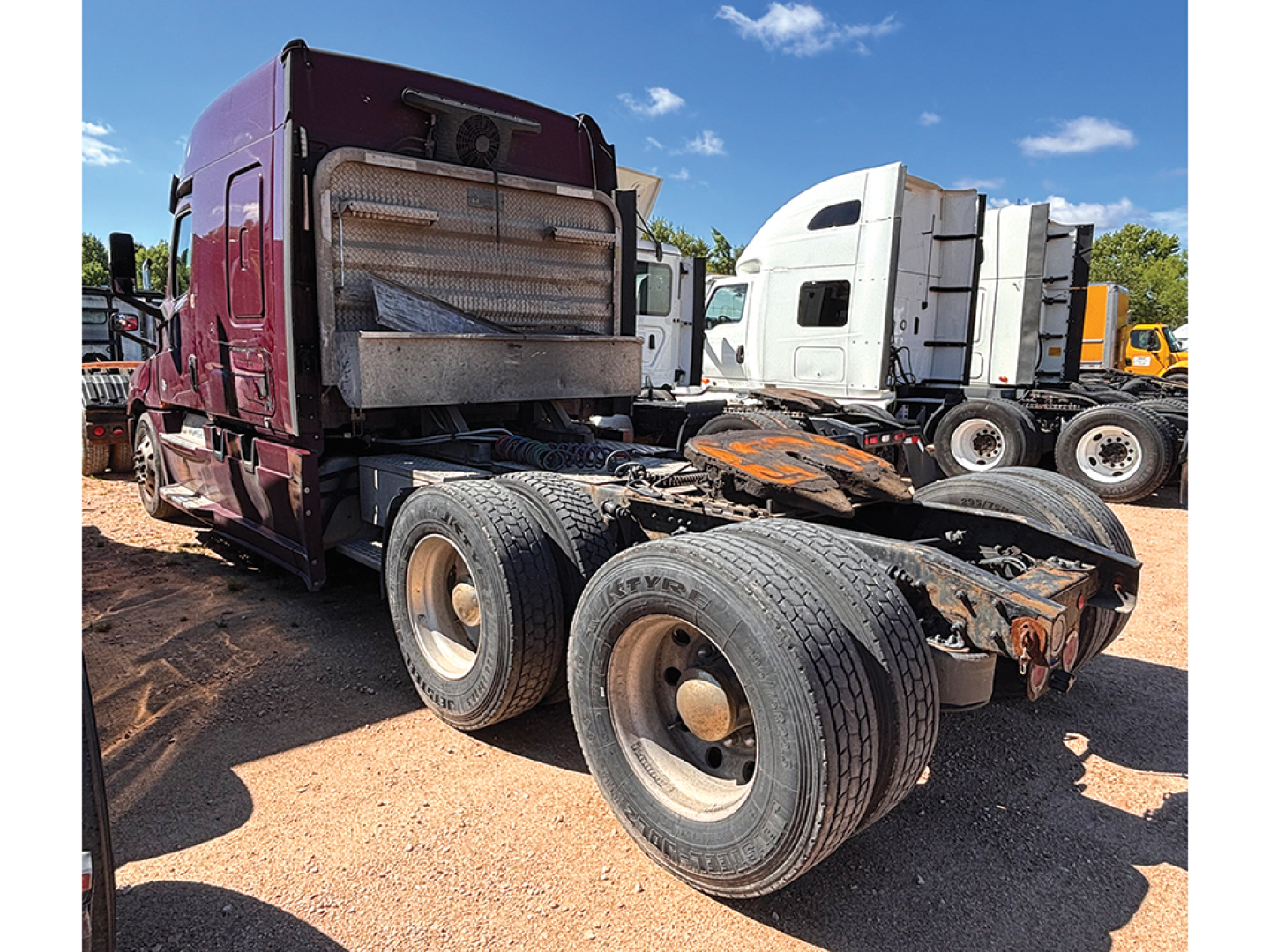 Truck chassis and cab of a 2019 Freightliner Cascadia126 from rear three-quarter view, with dual rear axles and fifth wheel missing, showing dirt and surrounding trucks.