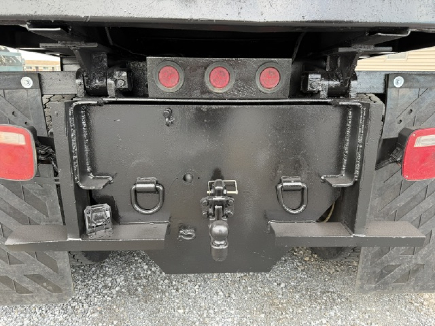 Truck rear showing a black steel toolbox with latches and a hitch, three red circular lights above, and mud flaps on a gravel surface.