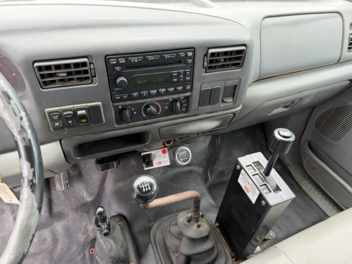 Truck cab interior with manual gear shift and array of controls, dashboard with radio, climate dials, and auxiliary equipment visible.