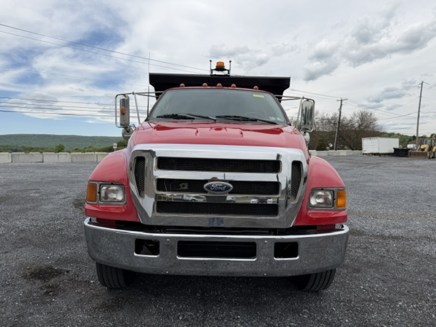 Front view of a red 2006 Ford F750 SD dump truck with chrome grille, bumper, and large side mirrors. Windshield shows faint text; truck is on gravel lot.