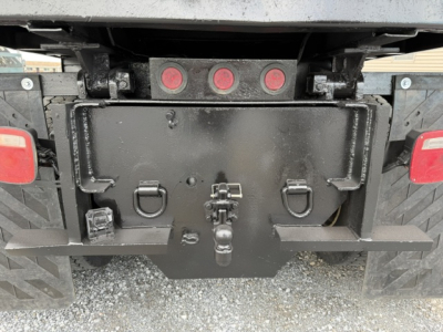 Truck rear showing a black steel toolbox with latches and a hitch, three red circular lights above, and mud flaps on a gravel surface.