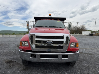 Front view of a red 2006 Ford F750 SD dump truck with chrome grille, bumper, and large side mirrors. Windshield shows faint text; truck is on gravel lot.