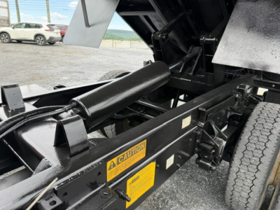 Ford F-750 SD truck chassis and hydraulic hoist mechanism under cargo bed with caution labels and tires in background.