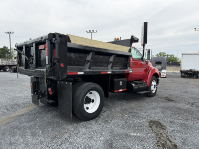 Ford F750 SD dump truck rear-right view showing black dump body with wooden bed, red cab, white rear wheels, and supporting hydraulic components.