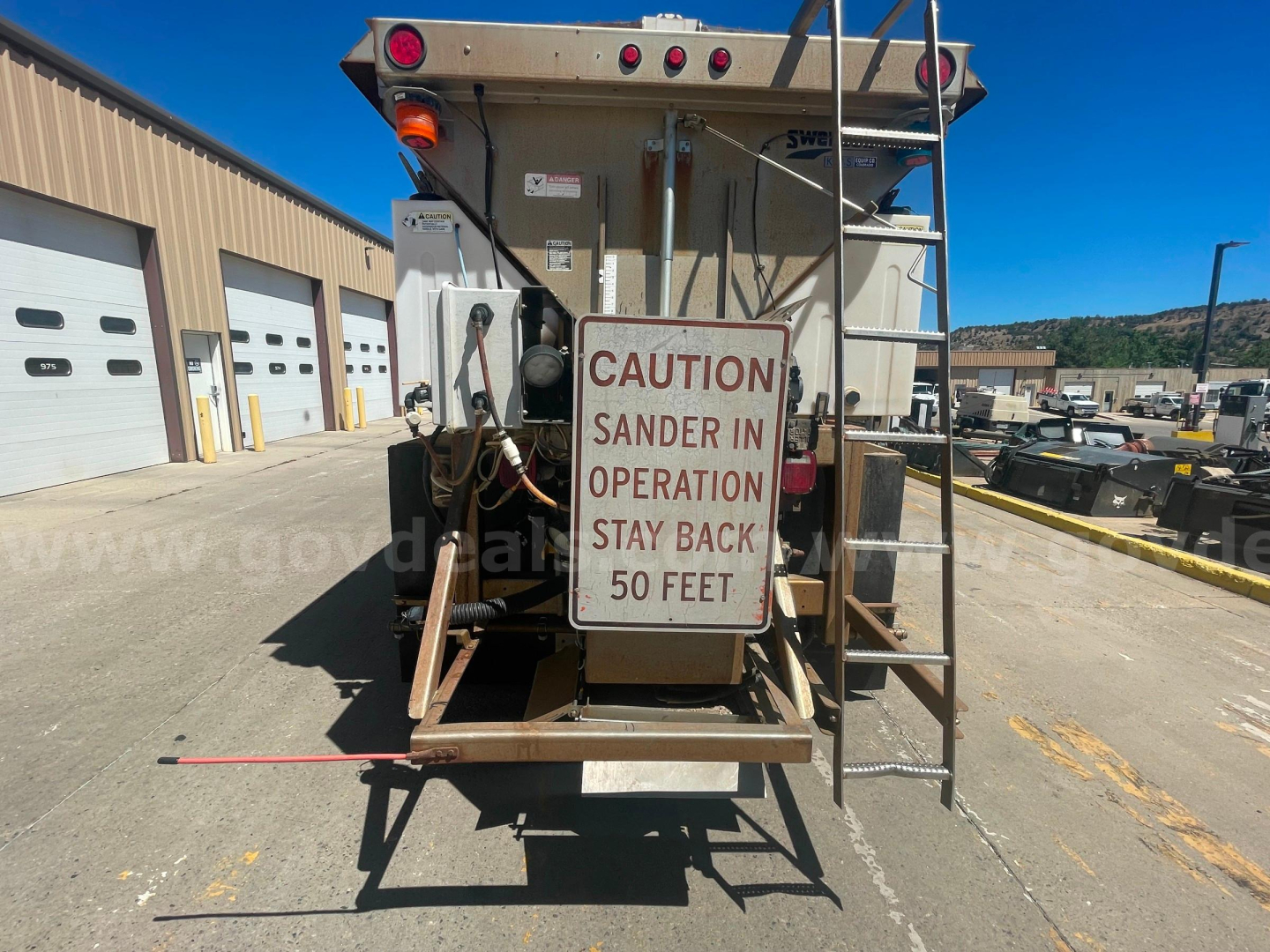 Dump truck rear showing caution sign: 'CAUTION SANDER IN OPERATION STAY BACK 50 FEET' on hopper; ladder and platform; equipment in yard.