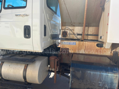 Truck cab and dump bed of an International 7400 with visible fuel tank, hydraulic components, and rust on frame; license plate obscured.
