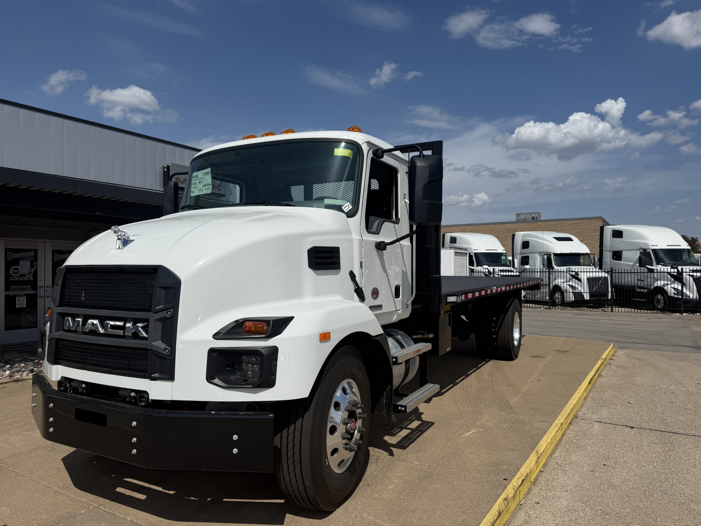 Mack MD6 flatbed truck in white with black bumper and dual rear wheels, parked in lot with other trucks in background.