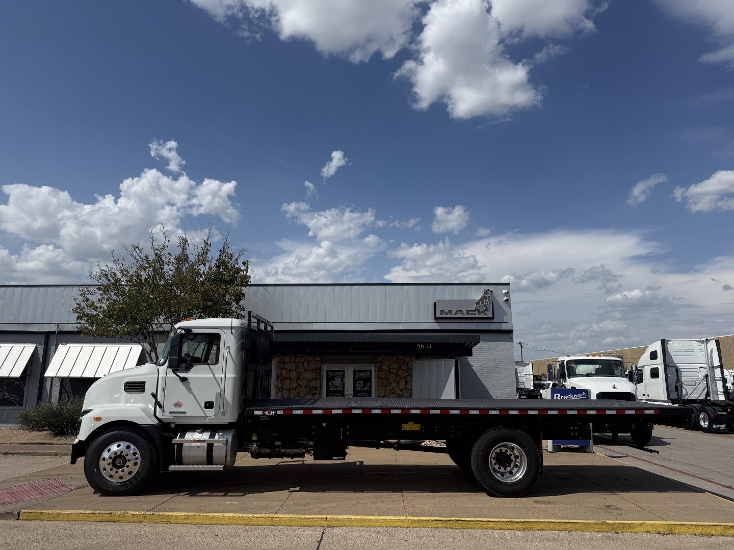 Flatbed truck side view showing Mack MD6 cab and long flatbed with black railing and red-and-white reflective tape.