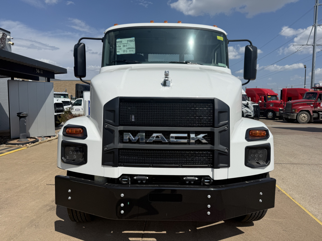Front view of a white 2026 Mack MD6 flatbed truck showing large black grille with MACK lettering, headlights, mirrors, and a protective front bumper.