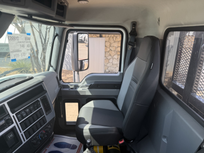 Interior cab view of a new 2026 Mack MD6 flatbed truck showing driver's seat and passenger seat, dashboard controls, side window, and door; no text legible besides interior features.