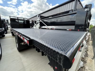 Flatbed truck bed of a 2026 Mack MD6 showing diamond plate steel surface, side rails, drop-down tailgate, and crew cab reflection in background.