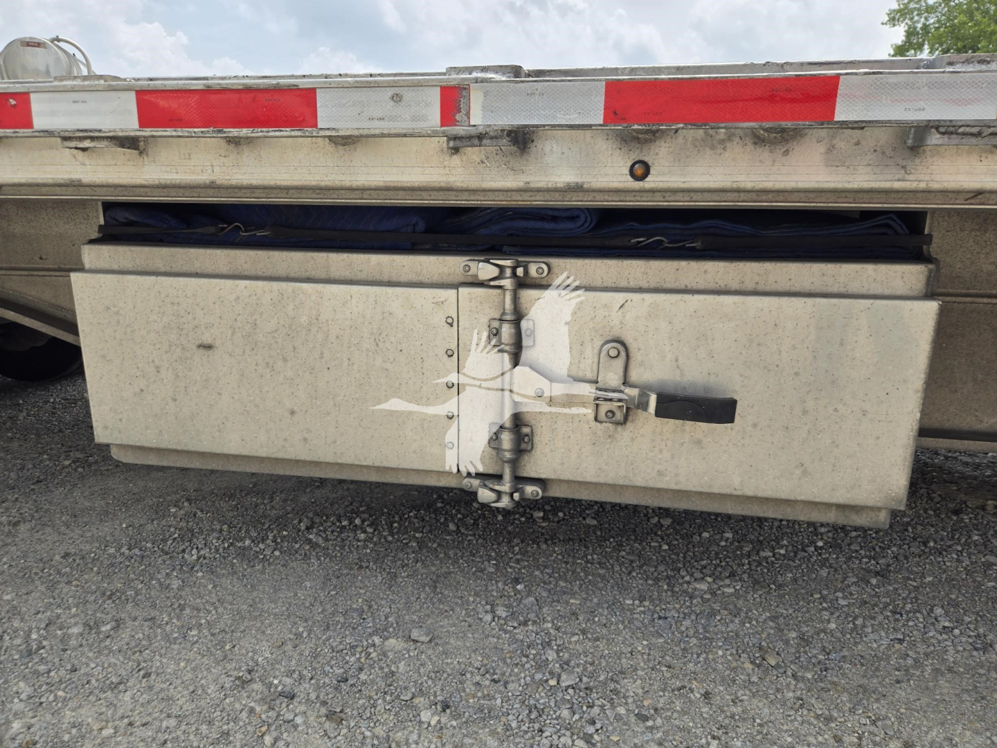 Flatbed trailer rear view showing a large rectangular aluminum toolbox mounted beneath the deck with latch and hinge hardware.