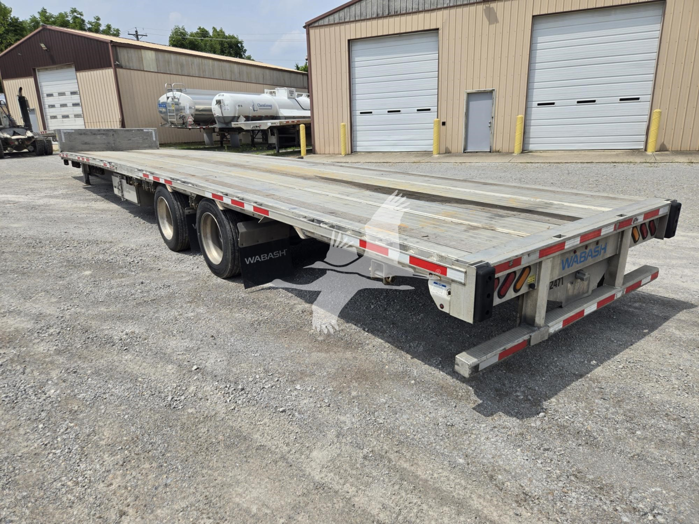 Trailer showing a long aluminum drop deck trailer with multiple axles and rear lights, parked on gravel near a warehouse.