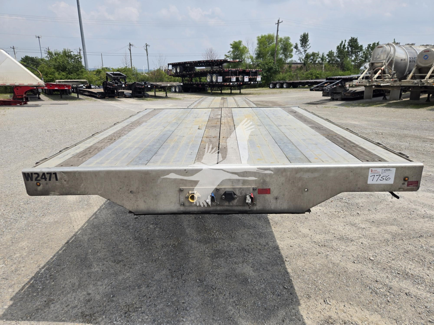 Trailer rear view of a used drop deck trailer with metal deck and uneven wear; two connection points and lighting visible on the bumper; serial/ID plate on left.