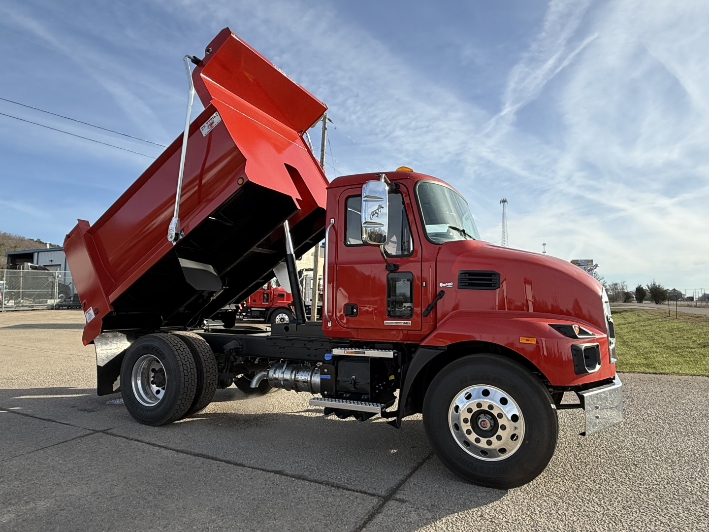 Dump truck with a raised red dump bed in a yard. Visible text on the cab includes no legible model text; general features include side mirror, front wheel, and steps.
