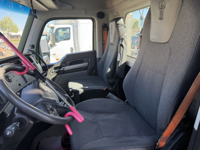 Interior front cab of a 2023 Kenworth T680 daycab with gray fabric seats, steering wheel, pink accents on handles, and dashboard visible.