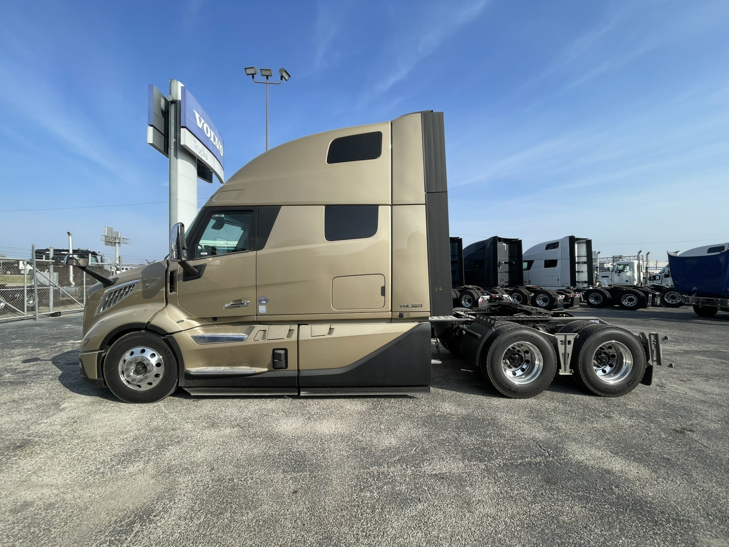 Volvo VNL cabin side view showing sleeper with tan paint, dual rear axles, and windows. No text clearly visible on the side, but a Volvo dealership sign in distance.