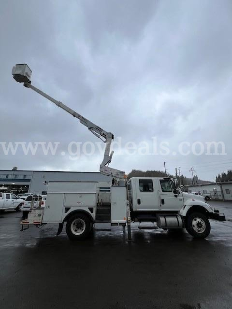 Truck side view of a 2006 International 7400 bucket truck with an extended aerial lift raised, utility body, white cab and chassis, in a parking lot.