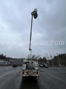 Bucket truck with extendable boom and bucket shown from rear, utility vehicle with equipment on bed, overcast sky.