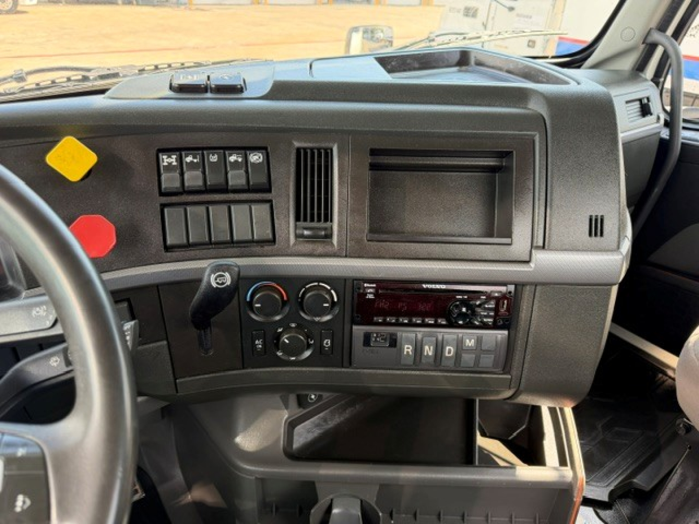 Volvo VNR64T300 dashboard view showing steering wheel, instrument cluster area, rows of toggle switches, square blank display, climate controls, and gear selector with R N D 3 2 1 labels.