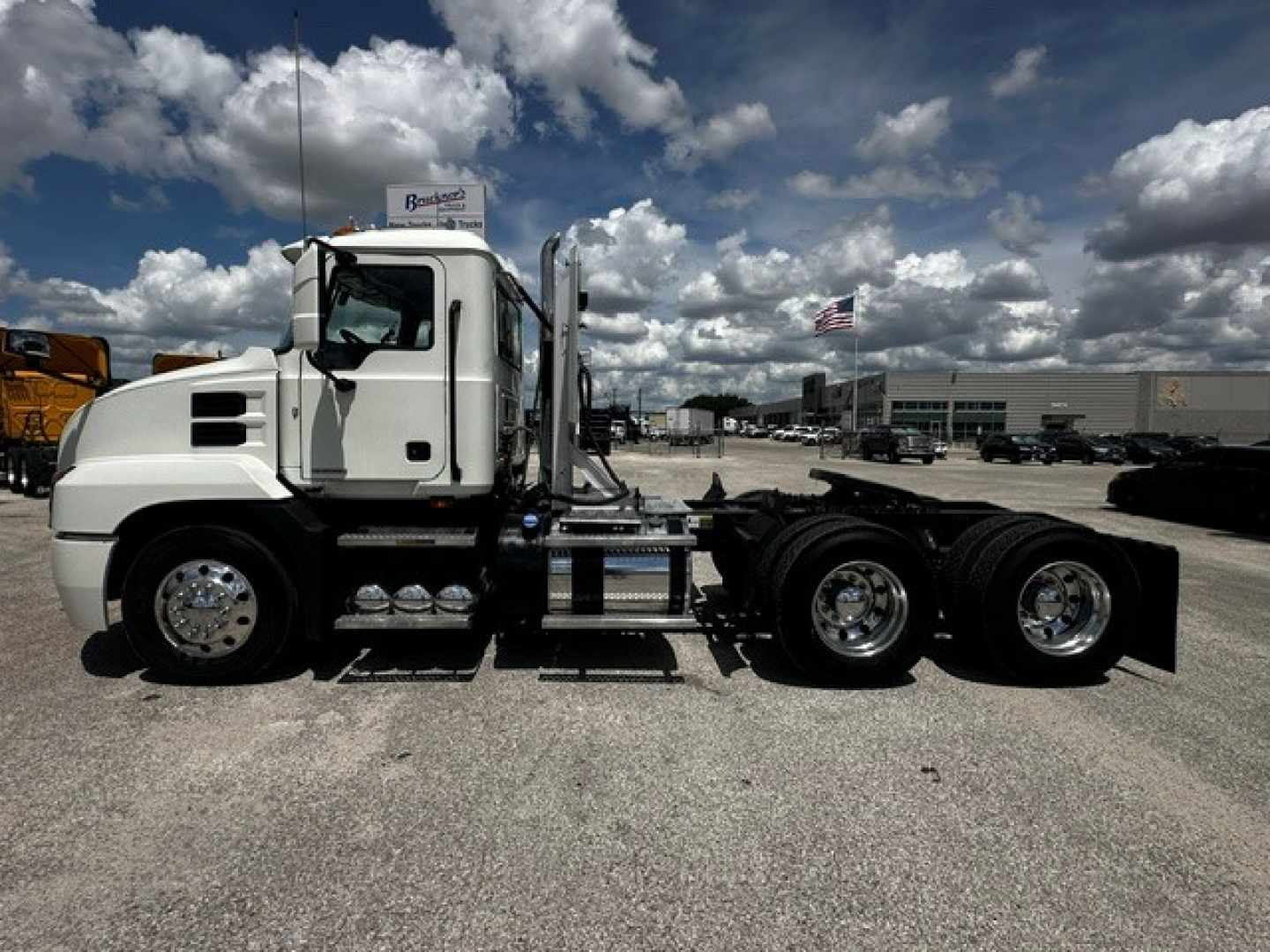 Truck chassis of a white 2019 Mack Anthem daycab, side view showing cab, fuel tanks, wheels, and rear axle without trailer, dealership lot in background.