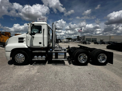 Truck chassis of a white 2019 Mack Anthem daycab, side view showing cab, fuel tanks, wheels, and rear axle without trailer, dealership lot in background.