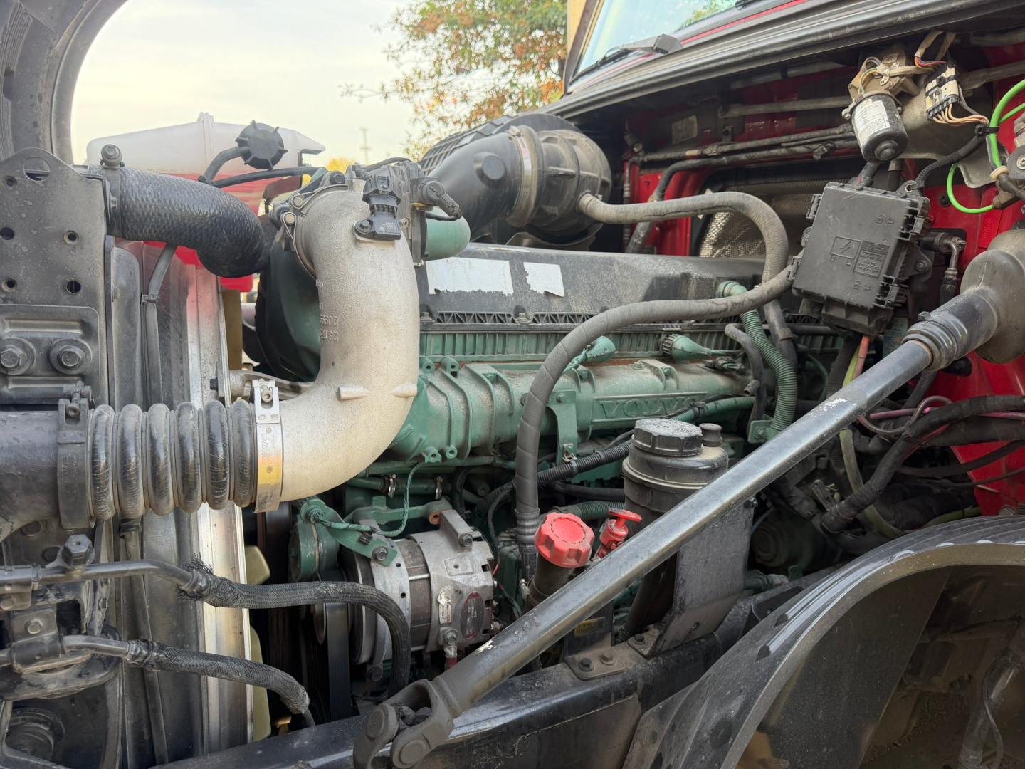 Truck engine bay showing Volvo diesel engine with green valve covers, radiator hoses, belts, and hoses in a red cab, label stickers and wiring harnesses visible.