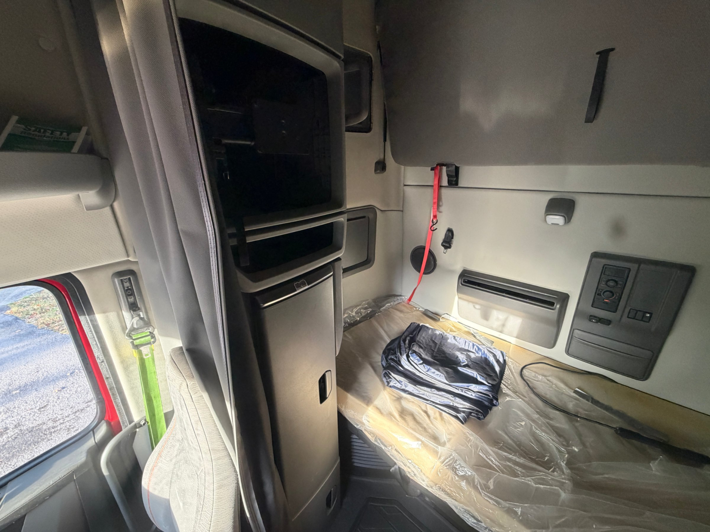 Cab interior of a Volvo VNL64T760 sleeper showing storage compartments and seat area with plastic covering and red battery jumper in view.