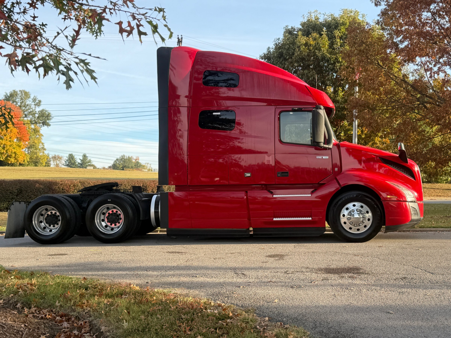 Side view of a red Volvo VNL64T760 semi-truck tractor with sleeper cab, three axles in back, black tires, chrome wheels, and trees in background.
