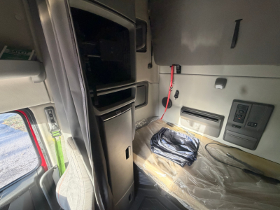 Cab interior of a Volvo VNL64T760 sleeper showing storage compartments and seat area with plastic covering and red battery jumper in view.