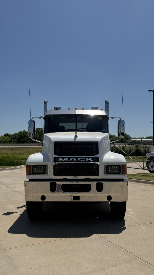 Front view of a white 2026 Mack PI64T day cab truck showing large grille, bumper, and Mack emblem. Clear sky, parking lot background.