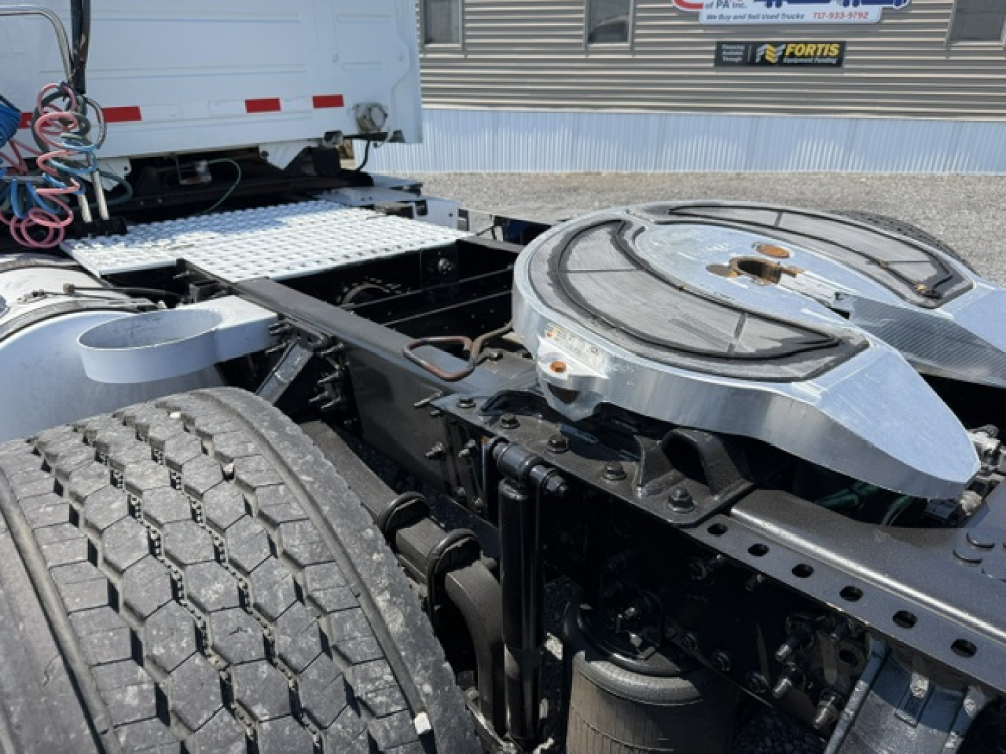 Volvo truck chassis in a yard with a circular aluminum fuel/def tank on the back, exposed frame, dual rear wheels, and a white cab visible in the background.