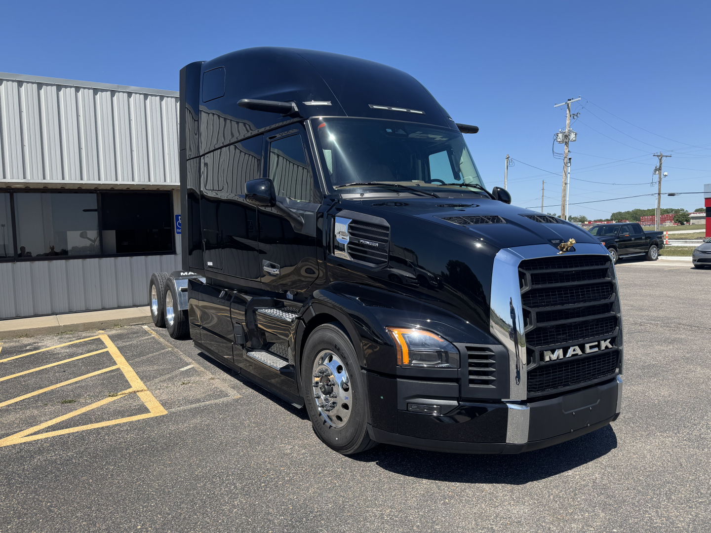 Truck cab side view of a black 2027 Mack PR64T76H sleeper, front grille with Mack logo, daytime sun, parking lot.