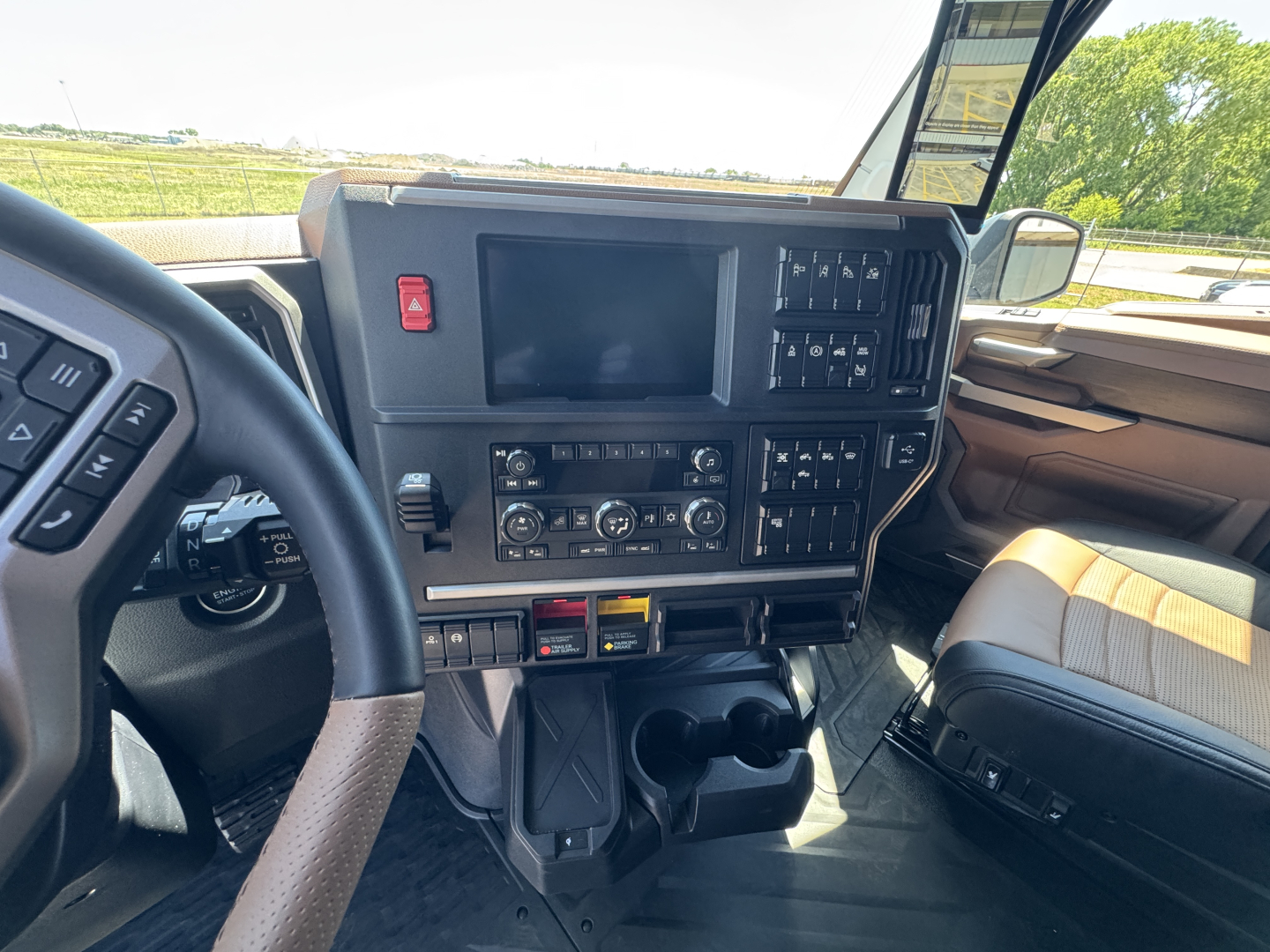 Mack truck cockpit with large central touchscreen, control panel, steering wheel, and driver seat visible. Dashboard displays various switches and gauges, dual cup holders, and sunlit interior.