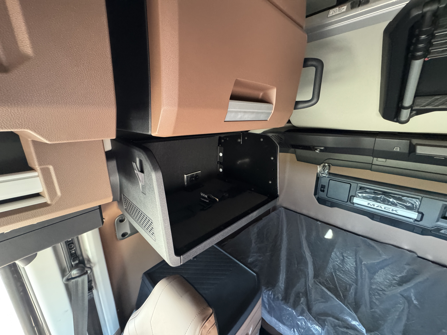 Truck cabin interior: open storage compartment under the dashboard in a Mack PR64T76H sleeper cab, showing a small empty bin and surrounding controls.