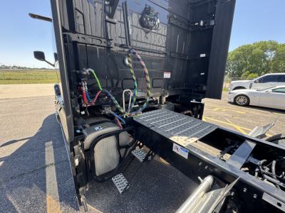 Truck cab area showing rear of tractor chassis with hydraulic lines and electrical wiring near sleeper, platforms, and battery box; Mack PR64T76H cab-forward configuration.