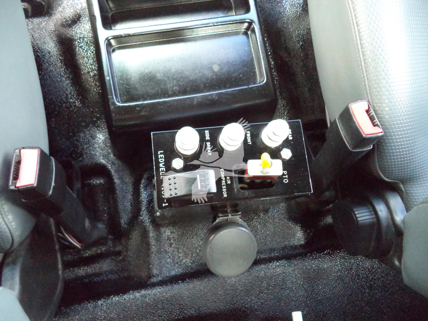 Ford F650 interior showing a control panel with three knobs and a yellow switch between two seats, cup holder in center, seat belts visible.