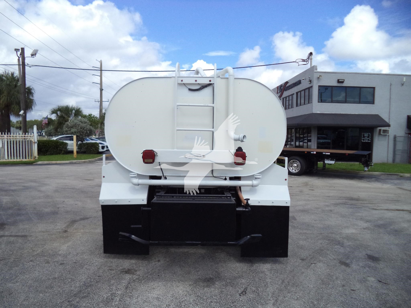 Back view of a white Ford F650/SD truck with a large cylindrical tank and rear platform, visible reflectors and ladder on tank, in a parking lot.