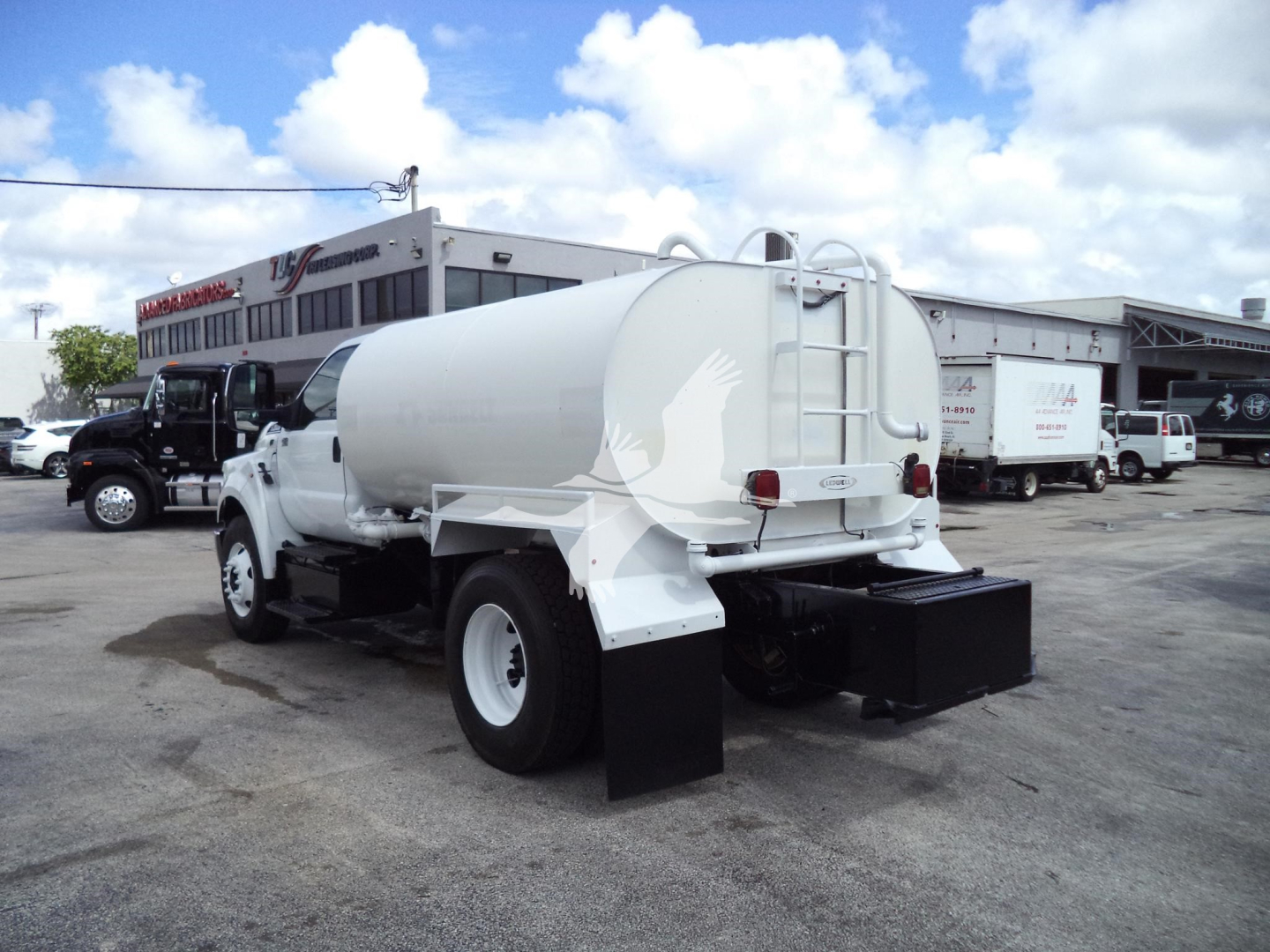 Ford F650 water truck with cylindrical tank and rear mud flaps, viewed from left-rear at a dealership lot.