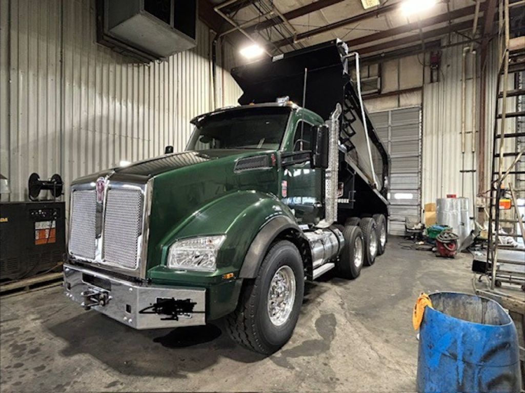 Kenworth T880 dump truck in a workshop, green cab with black dump bed raised, front chrome grille, badge, large tires, indoor industrial setting with equipment.