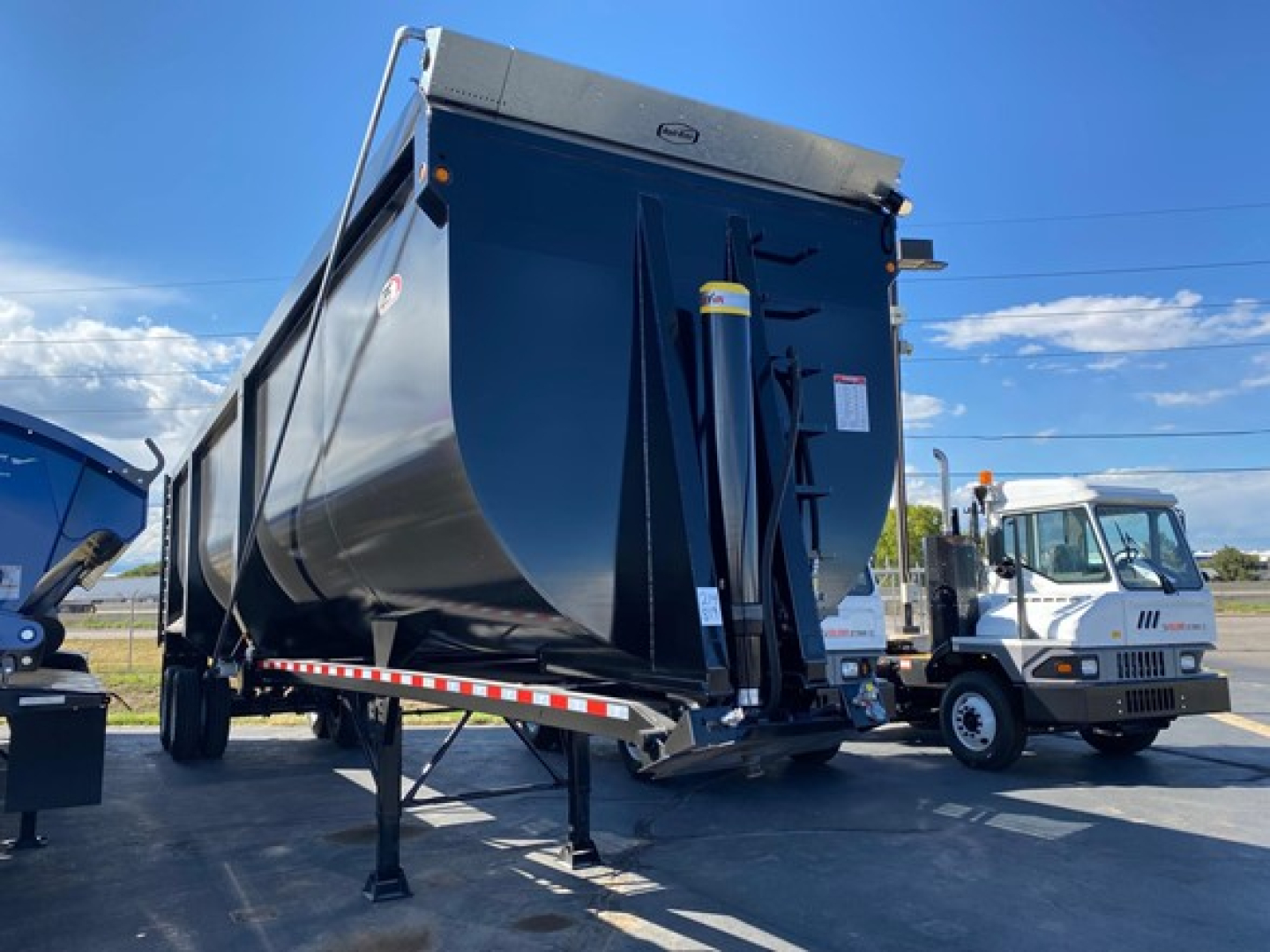 Dump trailer showing hoist mechanism and reinforced bed with front-mounted hitch and support legs; blue/gray color with reflective side rails and safety decals.
